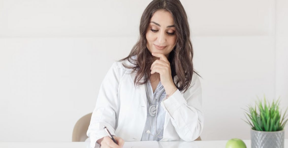 A female nutritionist in a lab coat writing notes with an apple and plant on the desk.