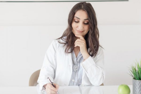 A female nutritionist in a lab coat writing notes with an apple and plant on the desk.